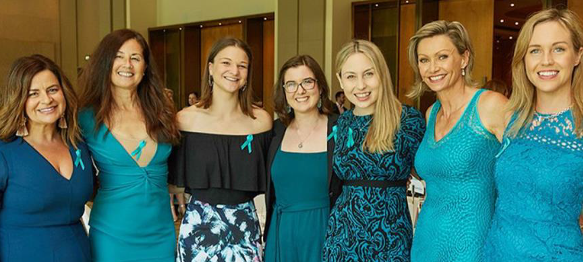 Seven women standing in a row, arms around each other, wearing blue outfits