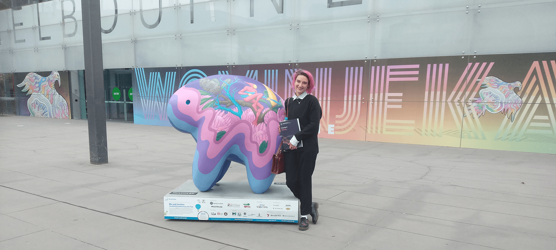 Nic with pink hair looking happy and confident. They're standing next to a colourful UooUoo sculpture outside Melbourne Museum