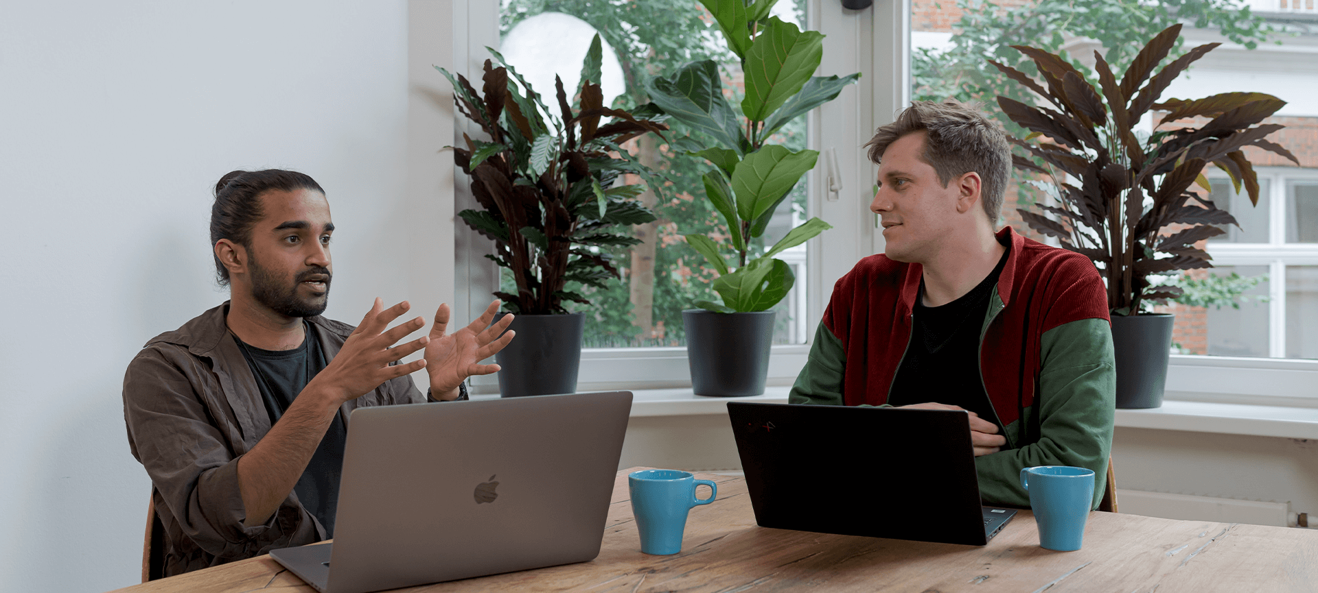 Two men sitting at a table talking with laptops and coffee