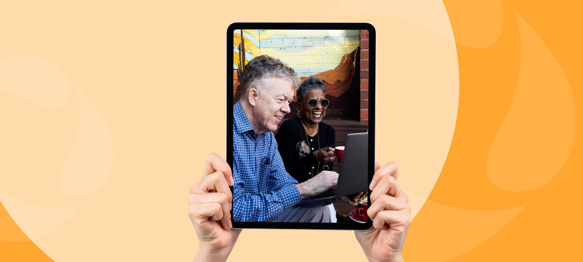 Hands holding a tablet displaying a photo of two smiling people at a laptop