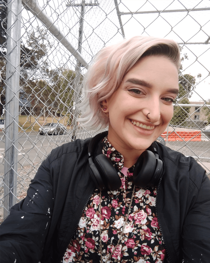 Nic with light pink hair and a nose ring looking happy and smiling at the camera. They have a colourful floral shirt on and headphones around their neck. Nic with light pink hair and a nose ring looking happy and smiling at the camera. They have a colourful floral shirt on and headphones around their neck.