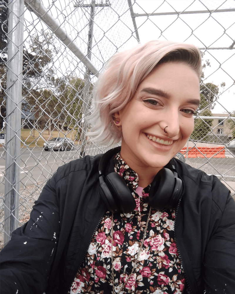 Nic with light pink hair and a nose ring looking happy and smiling at the camera. They have a colourful floral shirt on and headphones around their neck. 