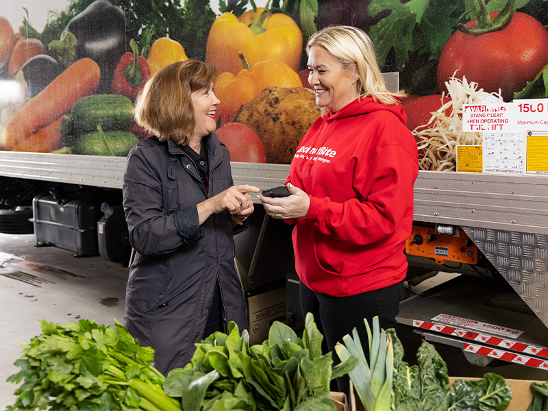 Zoe from SecondBite showing a woman Ask Izzy on a smartphone in front of a SecondBite truck